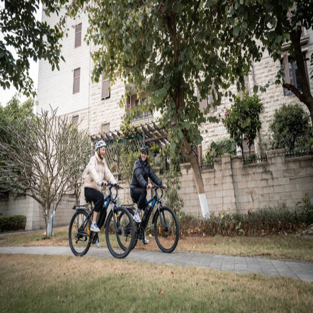 Two people riding bicycles on a path with trees and buildings in the background.
