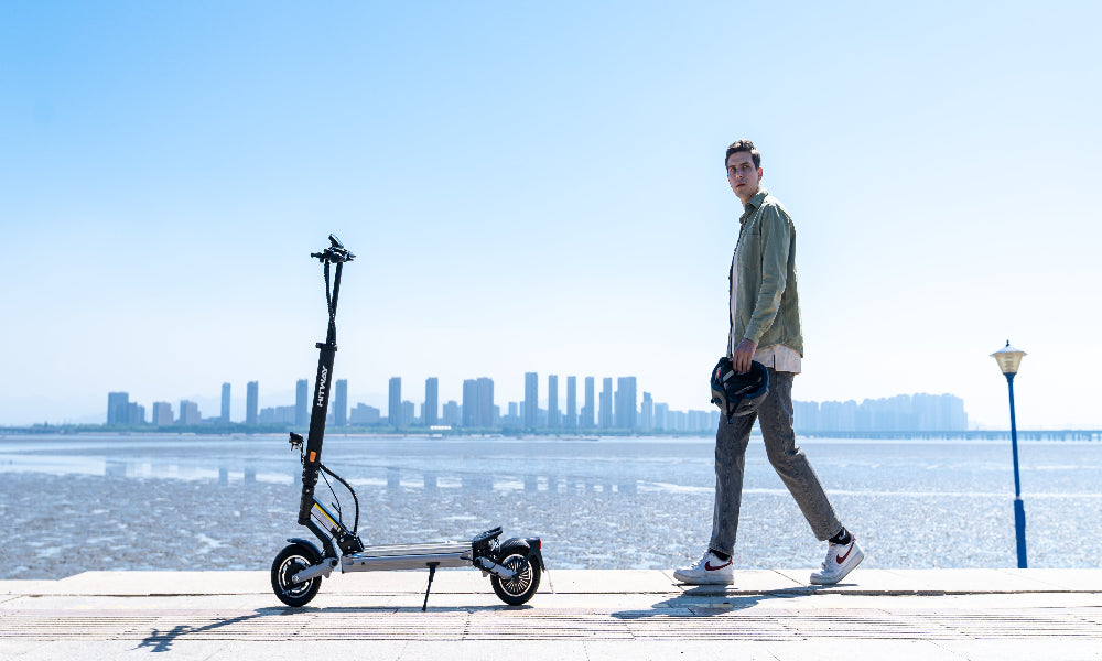 Man walking with an electric scooter by a waterfront with a city skyline in the background