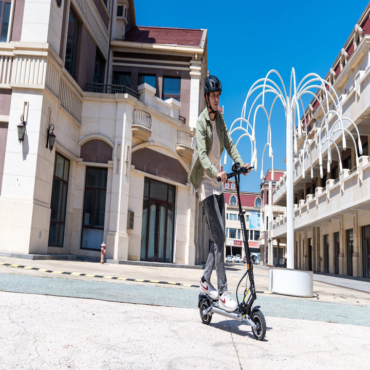 Person riding an electric scooter on a city street with buildings and a fountain in the background.