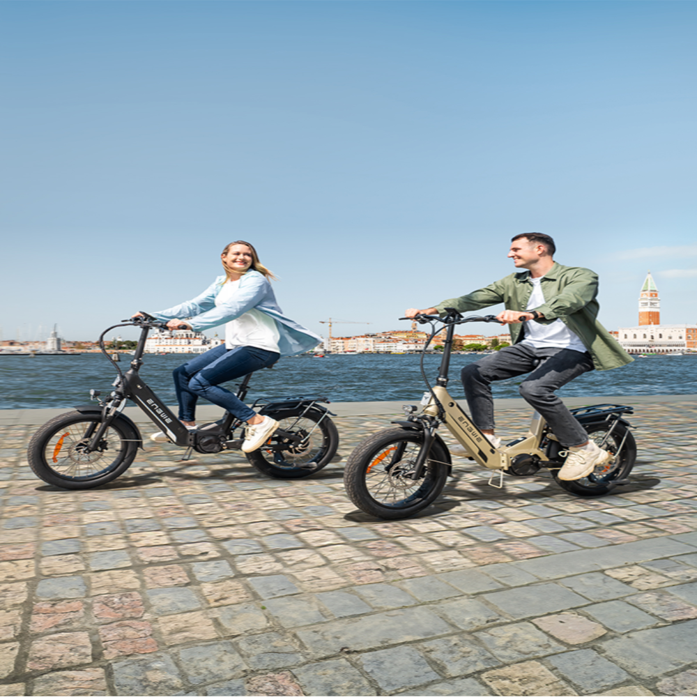 Two people riding electric bikes by a waterfront with a clear blue sky.