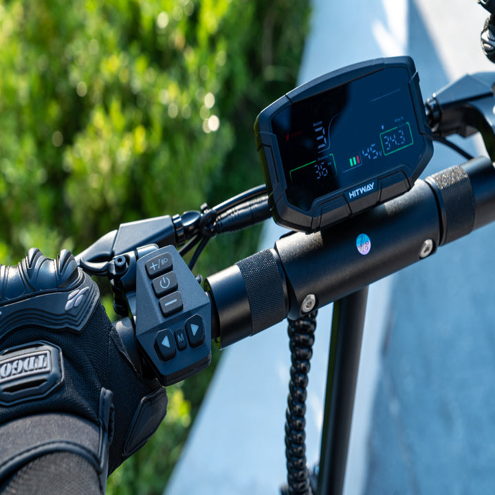 Close-up of a motorcycle handlebar with control panel and glove, blurred greenery in the background