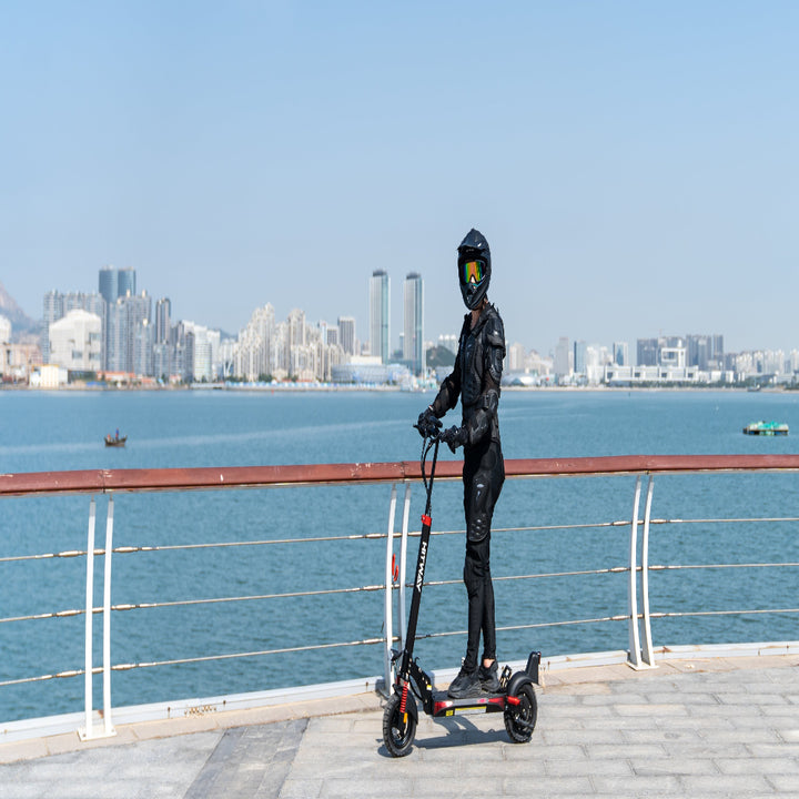 Person with a helmet and black outfit standing next to an electric scooter by a waterfront with city skyline in the background.