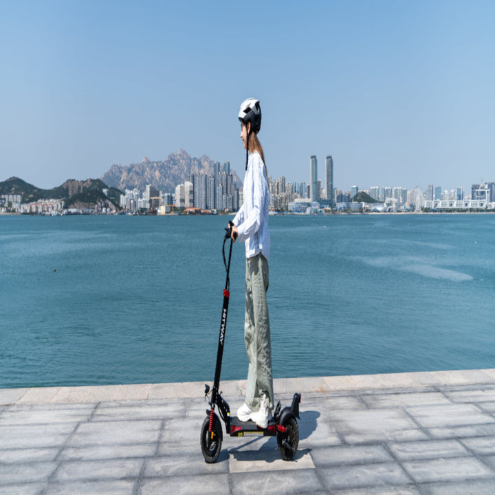 Person riding an electric scooter by a waterfront with a city skyline in the background