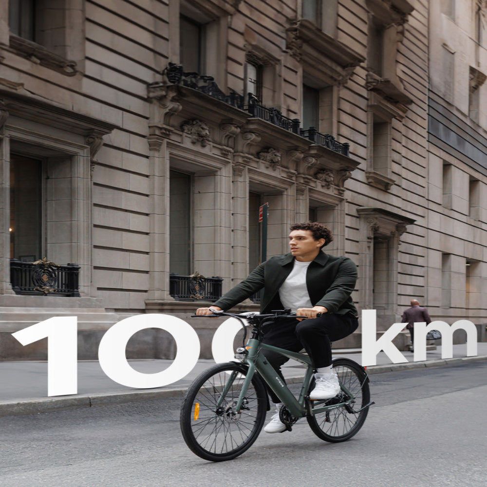 Man riding a bicycle on a city street with '100 km' text overlay