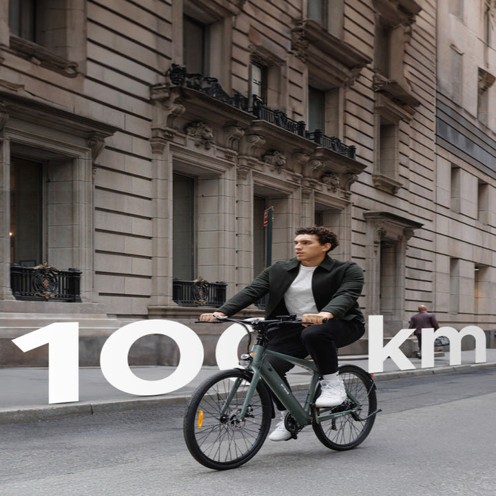 Man riding a bicycle on a city street with '100 km' text overlay