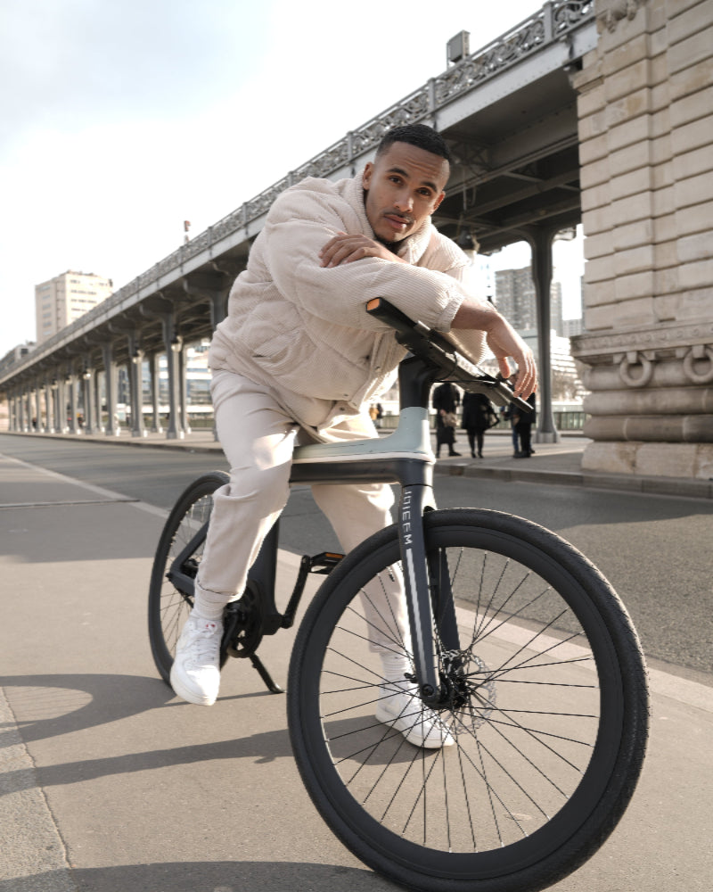 Man in a white outfit leaning on a black bicycle by a bridge